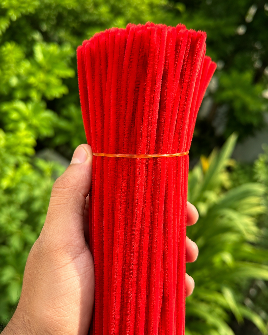 A hand holding a bundle of bright red fuzzy pipe cleaners tied with a thin orange string, with a blurred green garden background.